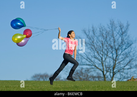 Ein junges Mädchen (11) läuft glücklich über grünen Rasen mit einer Reihe von bunten Luftballons hinter und blauer Himmel. Stockfoto