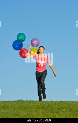 Ein junges Mädchen (11) läuft glücklich über grünen Rasen mit einer Reihe von bunten Luftballons hinter und blauer Himmel. Stockfoto