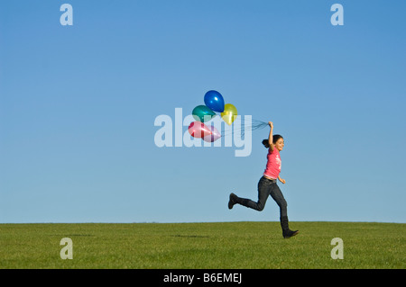 Ein junges Mädchen (11) läuft glücklich über grünen Rasen mit einer Reihe von bunten Luftballons hinter und blauer Himmel. Stockfoto