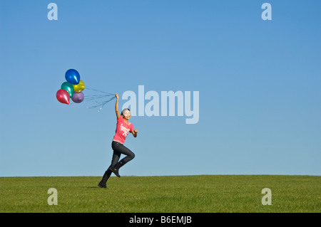 Ein junges Mädchen (11) läuft glücklich über grünen Rasen mit einer Reihe von bunten Luftballons hinter und blauer Himmel. Stockfoto