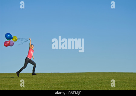 Ein junges Mädchen (11) läuft glücklich über grünen Rasen mit einer Reihe von bunten Luftballons hinter und blauer Himmel. Stockfoto