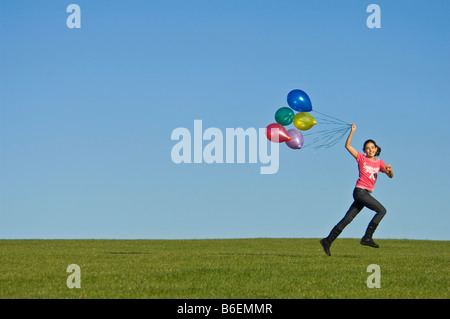 Ein junges Mädchen (11) läuft glücklich über grünen Rasen mit einer Reihe von bunten Luftballons hinter und blauer Himmel. Stockfoto