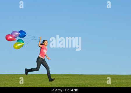 Ein Mädchen (11) läuft glücklich über grünen Rasen mit einer Reihe von bunten Luftballons hinter und blauer Himmel. Stockfoto