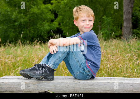 Ein Junge, sechs Jahre alt, sitzt auf einer Holzbank vor einem Laubwald Stockfoto