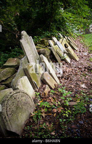 Alte ausrangierte, abgenutzte und gebrochene Grabsteinen gestapelt gegen eine Steinmauer mit dichtem Grün bewachsen. Stockfoto