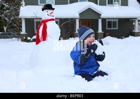 Kleiner Junge Essen Schnee mit Schneemann und Haus im Hintergrund Stockfoto