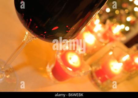 Abstraktes Konzept mit Glas gefüllt mit roten Wein und roten Votiv-Kerzen am Tisch vor Weihnachtsbeleuchtung und Baum angezündet. Stockfoto