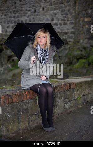 18 jährige Blondine mit schwarzen Regenschirm tragen Jacke und schwere Schal Blickkontakt auf Mäuerchen sitzen Stockfoto