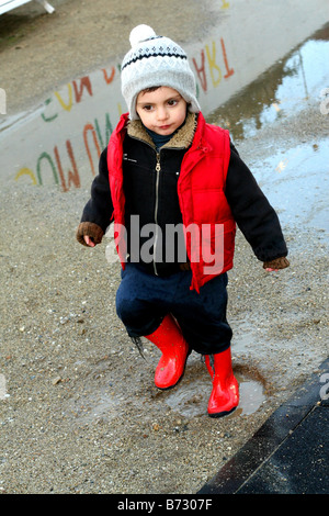 ein drei Jahre alter Junge Gummistiefel tragen und spielen in den Pfützen an einem regnerischen Tag Stockfoto