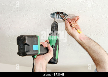 Eine Elektriker s Hände mit einem Bohrer, um eine Fan-Box in die Decke zu Schrauben, während er die Drähte aus dem Weg hält Stockfoto