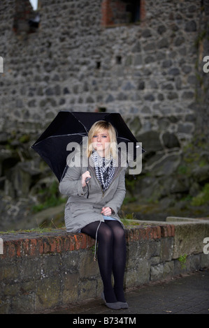 18 jährige Blondine hält Regenschirm tragen Jacke und schwere Schal sitzen auf niedrigen Mauer vor Carrickfergus castle Stockfoto