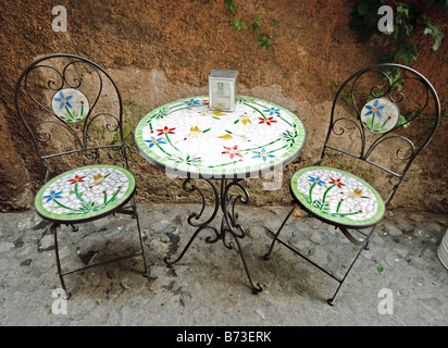 Sidewalk Cafe Rome Italy | ROME, Italy – Ein Bistrotisch mit Mosaikfliesen und zwei passende Stühle sitzen vor einer verwitterten ockerfarbenen Wand vor einem römischen Café, deren Blumenmuster aus gebrochenen Keramikfliesen zusammengesetzt sind. Schmiedeeiserne Bistro-Sets wie dieses sind gängige Elemente der Bürgersteige im historischen Zentrum Roms. Stockfoto
