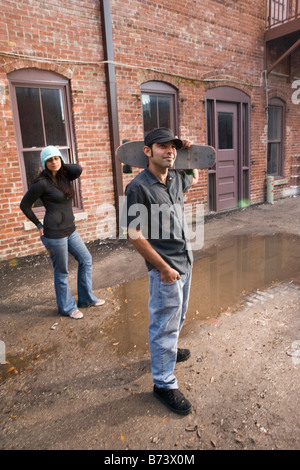 Junge Hispanic Mann in Gasse mit Skateboard, Frau im Hintergrund Stockfoto