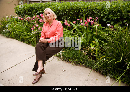 Reife Frau sitzend auf Stuhl auf Bürgersteig vor Blumen und Hecken Stockfoto