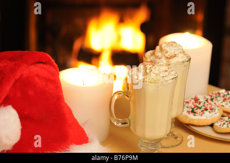 Glas Becher Weihnachten Eierlikör mit verzierten Plätzchen, Kerzen und Weihnachtsmütze mit Kamin brennen im Hintergrund Stockfoto