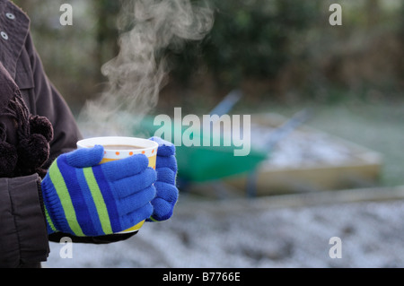 Lady Gärtner Hände in Handschuhe halten dampfenden Tasse Kaffee im Garten mit frostigen Garten im Hintergrund Januar Stockfoto