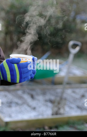 Lady Gärtner Hände in Handschuhe halten dampfenden Tasse Kaffee im Garten mit frostigen Garten im Hintergrund Januar Stockfoto