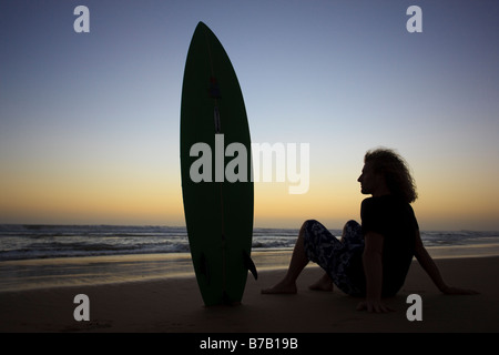 Ein stock Foto von einem jungen Mann mit seinem Surfbrett den Sonnenuntergang am Strand sitzen Stockfoto