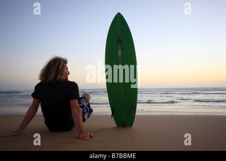 Ein stock Foto von einem jungen Mann mit seinem Surfbrett den Sonnenuntergang am Strand sitzen Stockfoto