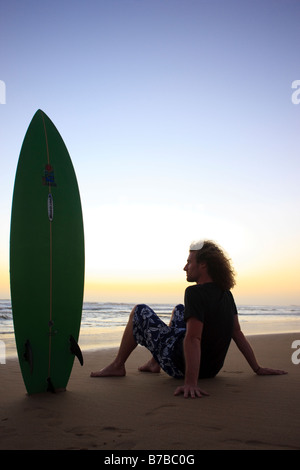 Ein stock Foto von einem jungen Mann mit seinem Surfbrett den Sonnenuntergang am Strand sitzen Stockfoto