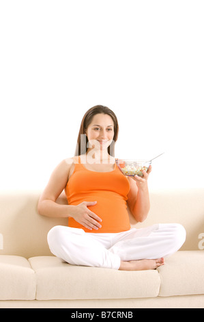 Schwangere junge Frau sitzt auf Sofa hält eine Schüssel Salat lächelnd Stockfoto