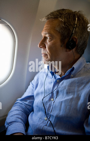 Ein männlicher Passagier Kopfhörer hören und schaut aus dem Fenster eines Flugzeugs Stockfoto