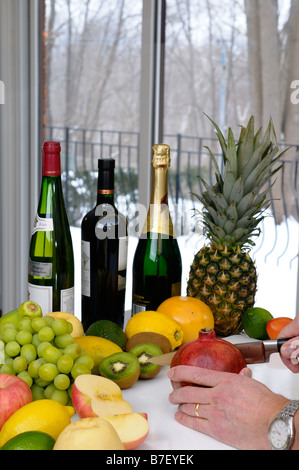 Man schneidet in einem Granatapfel, umgeben von Obst und Wein in einer Küche an einem Wintertag Stockfoto