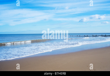 Sanft plätschernde Wellen plätschern auf saubere menschenleeren Sandstrand mit Buhne unter blassen blauen Himmel mit Wolken gestreift Stockfoto