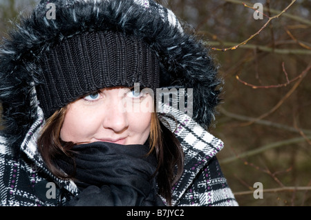 Junge Frau im Alter von 20-25 mit blauen Augen Lächeln auf den Lippen. Tragen Winterkleidung, wolligen Mütze, Schal und karierten Mantel. Im Wald Stockfoto