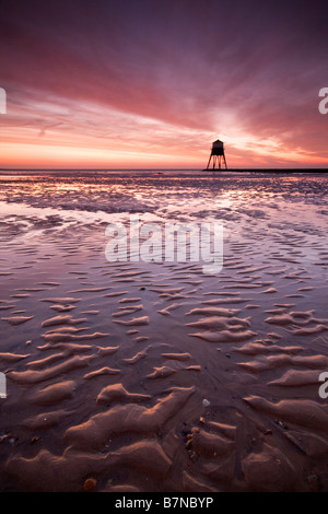 Dramatische Sonnenaufgang über den Strand bei Ebbe mit einem Leuchtturm in der Ferne Silhouette. Stockfoto