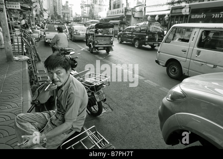 Mann Rauchen einer Zigarette am Straßenrand neben stark befahrenen entlang Thanon Yaowarat in Chinatown, Bangkok. Thailand Stockfoto