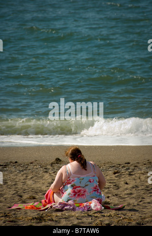 Eine weiße Frau, die von hinten am Strand sitzt und ein florales Badekostüm trägt, mit dem Meer in der Ferne. UK Stockfoto