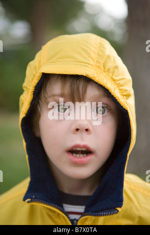 sechs Jahre alter Junge trägt einen gelben Regenmantel Kamera mit großen Augen starrte Stockfoto