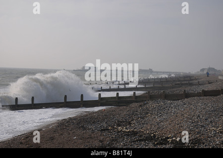 Eine Welle bricht über eine Buhne an einem einsamen Strand in Worthing während des Winters mit Worthing Pier im Hintergrund Stockfoto