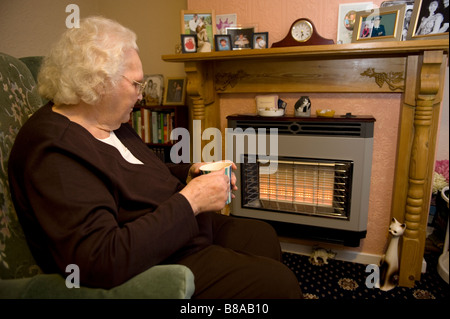 Eine ältere Frau hält warm vor ein elektrischer Kamin in ihrem Haus UK Stockfoto