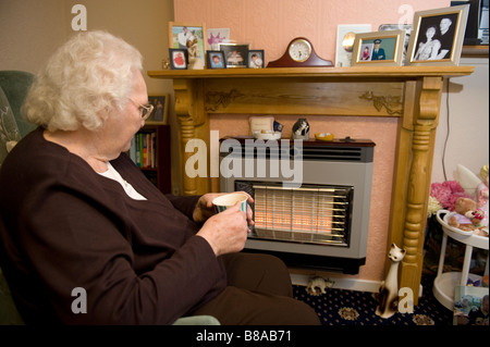 Eine ältere Frau hält warm vor ein elektrischer Kamin in ihrem Haus UK Stockfoto