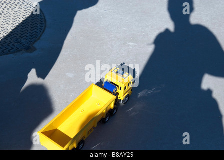 Schatten der stehend Leute fallen über ein Spielzeug aus Kunststoff gelb LKW auf Boden in der Nähe von Schachtabdeckung Stockfoto