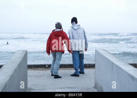 eine Rückansicht von zwei älteren Menschen Hand in Hand und stehen auf einer Strandpromenade mit Blick auf das Meer an einem stürmischen Tag Stockfoto