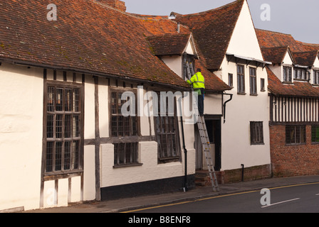 Fensterputzer am Arbeitsplatz auf eine Leiter trägt einen hohe Sichtbarkeit Mantel in Lavenham, Suffolk, Uk Stockfoto