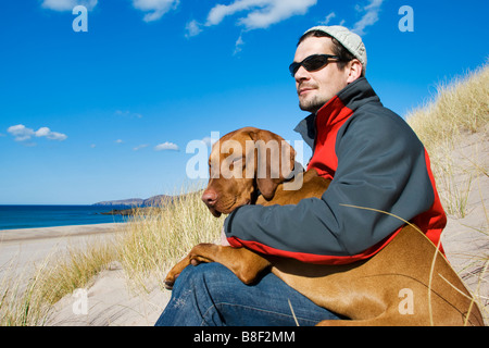 Mann saß mit Magyar Vizsla Hund auf seinem Schoß auf die isolierte uk Strand Sandwood Bay in Schottland Stockfoto