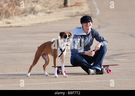 Ein hübsches Mädchen mit ihrem Boxer Hund an der Leine ruht auf ihrem Skateboard. Stockfoto