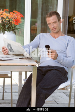 Mitte erwachsenen Mann sitzt auf der Terrasse halten eines Handys während des Lesens einer Zeitungs Stockfoto