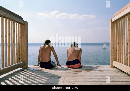 Zwei Jungs sitzen nebeneinander am Rande einer Fußgängerbrücke - schwimmen - Freundschaft - Freizeit - Jugend - See Stockfoto