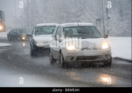 Autos fahren langsam auf einer verschneiten Straße im Winter in England mit Scheinwerfern mit Schnee bedeckt Stockfoto