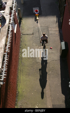 Ein kleiner Junge, ziehen einen Wagen auf seinem Fahrrad durch eine Siedlung mit einem Sicherheitszaun zu seiner linken an einem Sommernachmittag. Stockfoto