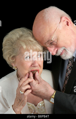 Ein hübscher senior Mann romantisch Fütterung Pralinen, seine schöne Frau schwarzen Hintergrund Stockfoto