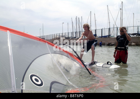 junger Mann Junge auf einem Surfbrett Surfkurse einzuräumen. Der junge versucht, heben Sie das Segel aus dem Wasser, Spanien, Balearen, Majo Stockfoto