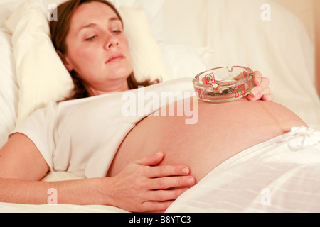 Nahaufnahme von einer vollen gebrauchte Aschenbecher auf den wachsenden Bauch Bump der eine junge schwangere Frau im dritten Trimester der Schwangerschaft Stockfoto