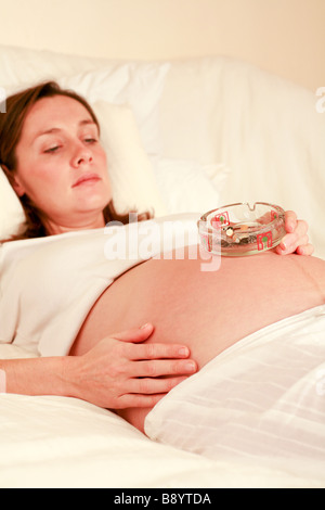 Nahaufnahme von einer vollen gebrauchte Aschenbecher auf den wachsenden Bauch Bump der eine junge schwangere Frau im dritten Trimester der Schwangerschaft Stockfoto