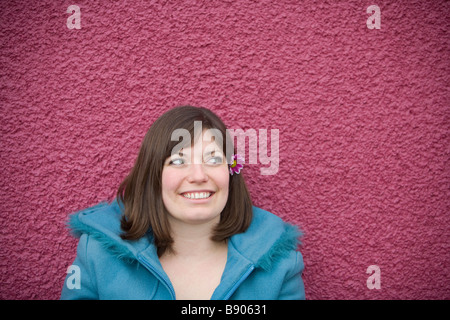 Eine junge Brünette Frau trägt eine rosa Blume im Haar und eine blaue Jacke im Freien vor einer rosa Wand. Stockfoto